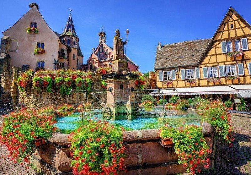 A village fountain ringed with geraniums below a church and painted houses